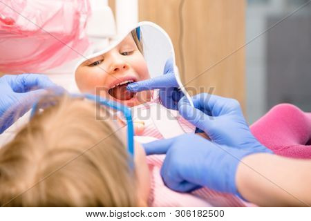 Dentist showing to little girl cured tooth in pediatric dental clinic. Child is sitting in a dental chair inspecting her teeth looking thru tooth-shaped mirror
