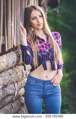 Cowgirl In Jeans And Checkered Shirt Standing By The Wooden Wall