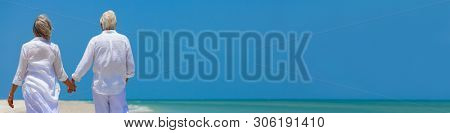 Panora rear view of happy successful retired senior man and woman couple walking and holding hands on a deserted tropical beach with bright clear blue sky panoramic web banner