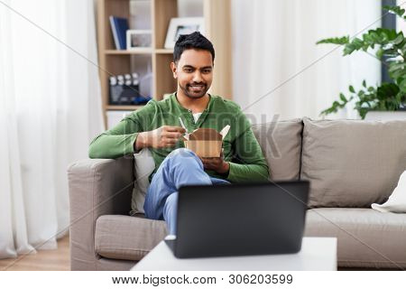 consumption and people concept - smiling indian man eating takeaway food and watching something on laptop computer at home