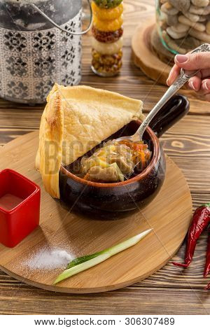 Female Hand Holding Spoon With Russian Cabbage Soup Shchi With Beef In Clay Pot On Wooden Table Side