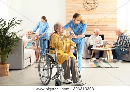 Nurse Giving Glass Of Water To Elderly Woman In Wheelchair At Retirement Home. Assisting Senior Peop