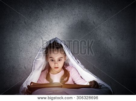 Engrossed Little Girl Reading Book In Bed Before Going To Sleep. Pretty Kid With Flashlight Hiding U