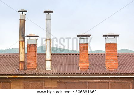 Brick Chimneys And Tin Ventilation Pipes On The Roof Of A Wooden Hut On A Background Of Green Hills 