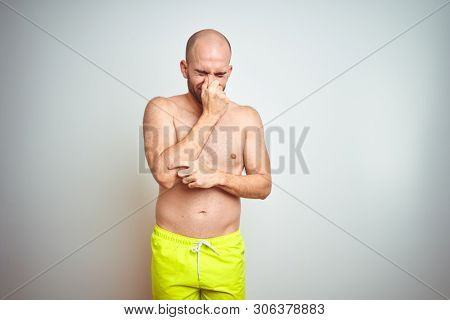Young shirtless man on vacation wearing yellow swimwear over isolated background smelling something stinky and disgusting, intolerable smell, holding breath with fingers on nose. Bad smells concept.