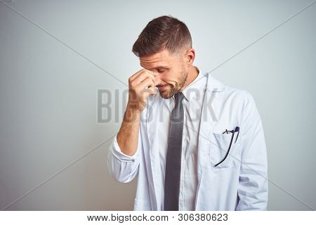 Young handsome doctor man wearing white profressional coat over isolated background tired rubbing nose and eyes feeling fatigue and headache. Stress and frustration concept.