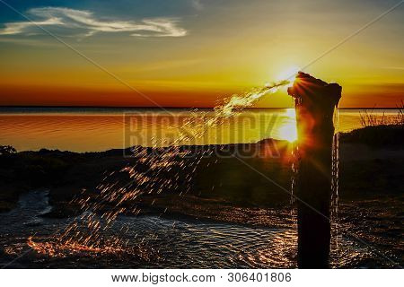 A Jet Of Water From A Well Of Golden Color Against The Sunset Over The Lake