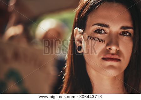 Close Up Of Young Activist With Word Freedom Written On Her Face Looking At Camera While Protesting 