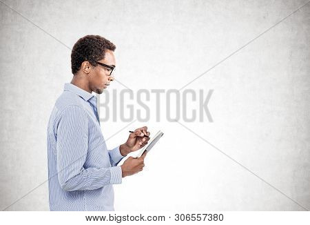 Side Portrait Of Serious Young African American Man In Blue Shirt And Glasses Taking Notes Standing 