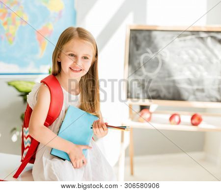 Little smiling blond girl standing near blackbord with red backpack and holding a notebook with pencils in the sunshine school classroom