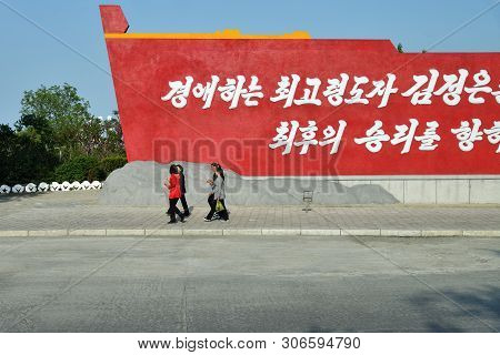 Wonsan, North Korea - May 3, 2019: Propaganda Banner And Groups Locals Girl Walking Down The Street 
