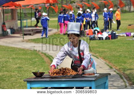 Pyongyang, North Korea - May 1, 2019: Female Seller Of Roast Meat Shown In The Taesongsan Funfair. I