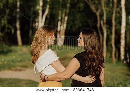 Young Women Different Build With Long Hair Are Embracing On The Green Field In The Evening. Friendsh