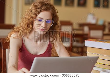 Indoor Shot Of Concentrated Beautiful Lady Student Working With Laptop At Library, Making Notes On H