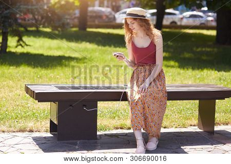 Image Of Young Girl Charges Mobile Phone Via Usb Outdoors, Female Sitting On Bench With Solar Panel 