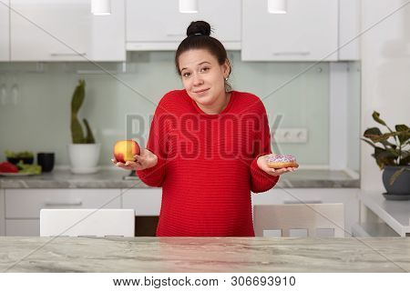 Indoor Shot Of Young Pregnant Woman With Apple And Tasty Cake In Her Hands, Decides What To Eat, Att