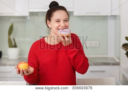 Pregnant Woman Eating Sweet Cake And Holding Apple In Another Hand, Stands Wearing Red Casual Sweate