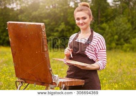 Outdoor Shot Of Young Woman Artist Standing With Sketchbook Against Meadow Background, Holding Brush