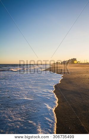 Waves Of White Sea Foam Washing Up On A Beach In Evening Light.