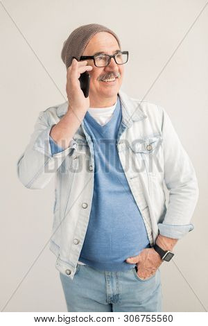 Aged cheerful man in casual pullover, jacket, blue jeans and beanie calling by smartphone while standing by white background