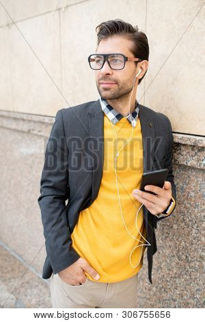 Contemporary young mobile man in smart casual and earphones listening to music while standing by wall of modern building