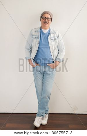 Happy mature man in jeans, pullover, jacket, beanie and sneakers leaning over white wall in studio while looking at you