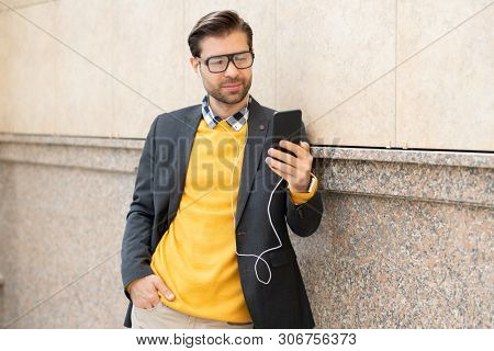 Stylish young man in smart casual and earphones standing by wall of building and making selfie while listening to music