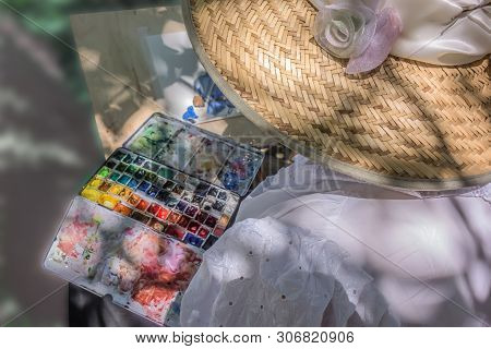 Woman Artist In A Beautiful Straw Hat, Decorated With White Feathers And Flowers, Sits In Front Of T