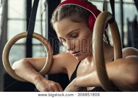 Closeup face of young tired athlete listening to music over headphones while relaxing on gymnastic rings. Exhausted woman resting on gymnastic rings listening to motivational music with headset.