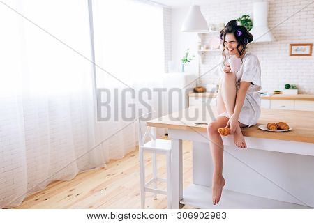 Cheerful Careless Young Woman With Curlers In Haor. Sitting On Table In Kitchen And Laughing. Hold C