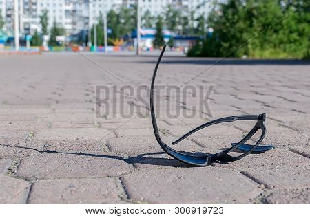 Broken Sun Glasses Lie On A Stone Tiled Walkway In The City Park Against The Background Of Residenti