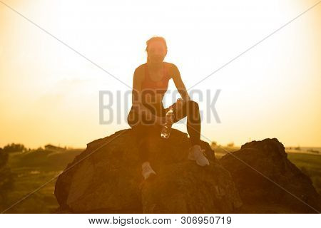 Young Woman Resting and Drinking Water after Trail Running Outdoor. Workout Exercise at Hot Beautiful Summer Sunset. Sport and Healthy Active Lifesyle Concept.