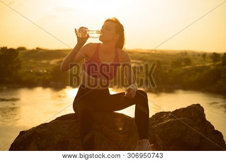 Young Woman Resting and Drinking Water after Trail Running Outdoor. Workout Exercise at Hot Beautiful Summer Sunset. Sport and Healthy Active Lifesyle Concept.