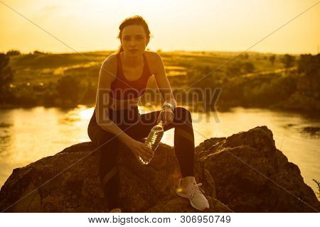 Young Woman Resting and Drinking Water after Trail Running Outdoor. Workout Exercise at Hot Beautiful Summer Sunset. Sport and Healthy Active Lifesyle Concept.