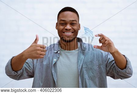 End Of Coronavirus Pandemic. Happy Black Guy Taking Off Face Mask Gesturing Thumbs Up Over White Bac