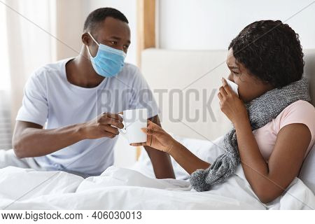 Black Husband In Protective Face Mask Taking Care Of His Sick Wife, Giving Hot Drink, Bedroom Interi