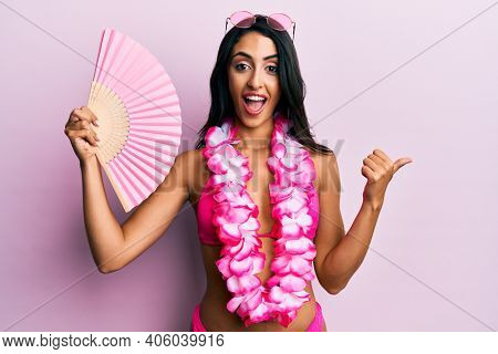 Beautiful hispanic woman wearing bikini holding handfan pointing thumb up to the side smiling happy with open mouth 