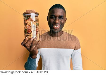 Young african american man holding jar with farfalle pasta looking positive and happy standing and smiling with a confident smile showing teeth 