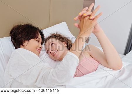 Beautiful Young Lesbian Women Lying In Bed, Holding Hands And Looking At Each Other