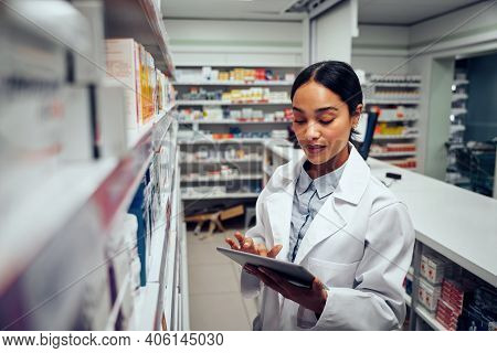 Young Female Pharmacist Checking Inventory Of Medicines In Pharmacy Using Digital Tablet Wearing Lab