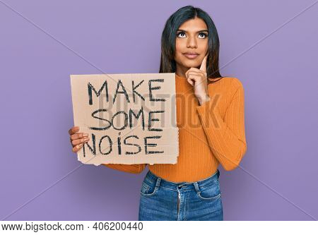 Young latin transsexual transgender woman holding make some noise banner serious face thinking about question with hand on chin, thoughtful about confusing idea 