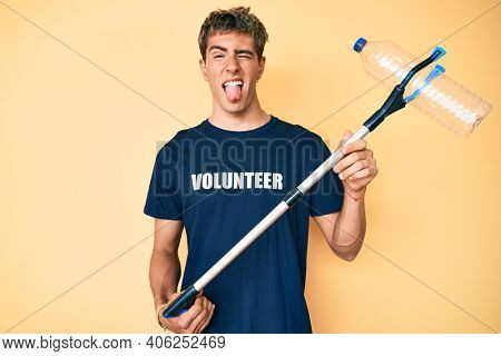 Young handsome man recycling plastic bottle holding litter pick up tool sticking tongue out happy with funny expression. 