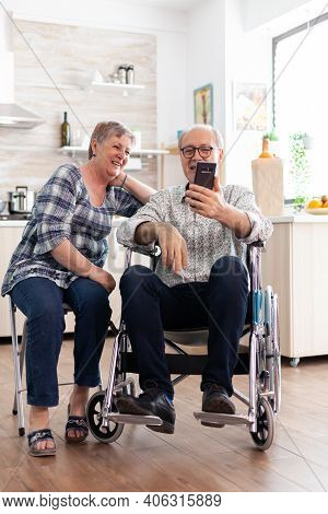 Smiling Senior Woman And Her Disabled Husband In Wheelchair Using Smartphone In Kitchen, Surfing On 