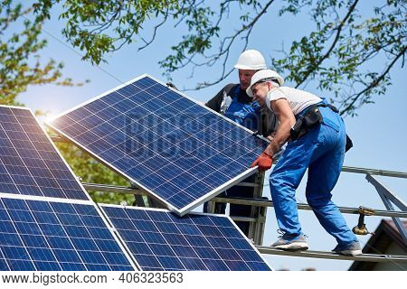 Two Young Technicians Mounting Heavy Solar Photo Voltaic Panel On Tall Steel Platform On Green Tree 