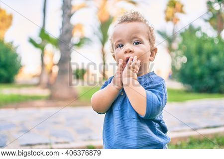 Sad little boy putting fingers on mouth touching gums because toothache at the park on a sunny day. Beautiful blonde hair male toddler in pain for new baby teeth outdoors