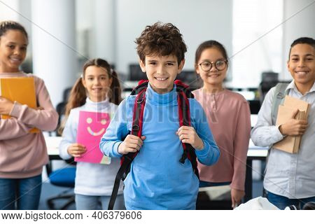 Return Back To School. Smiling Small Boy Wearing Backpack Posing In Classroom, Diverse Group Of Happ