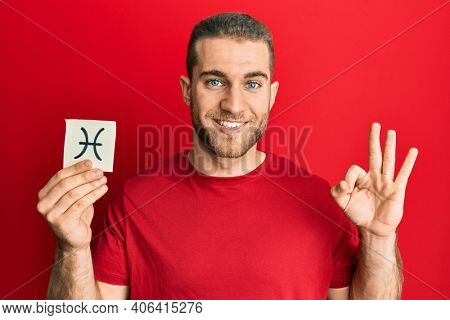 Young caucasian man holding paper with pisces zodiac sign doing ok sign with fingers, smiling friendly gesturing excellent symbol 