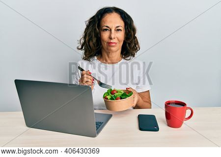Beautiful middle age woman working at the office eating healthy salad relaxed with serious expression on face. simple and natural looking at the camera. 