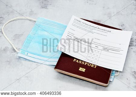 Coronavirus Vaccination Record Card, Biometric Passport And Blue Medical Mask On Light Gray Desk. Co