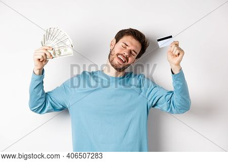 Handsome Man Dancing With Money And Plastic Credit Card, Standing In Casual Clothes Over White Backg